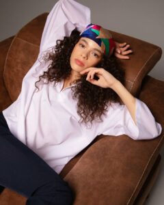 Femme avec des cheveux bouclés portant un foulard coloré, allongée sur un canapé en cuir marron.