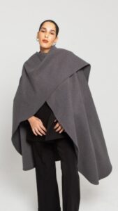 Femme portant un poncho gris sur un haut noir et un pantalon noir, avec une expression neutre.