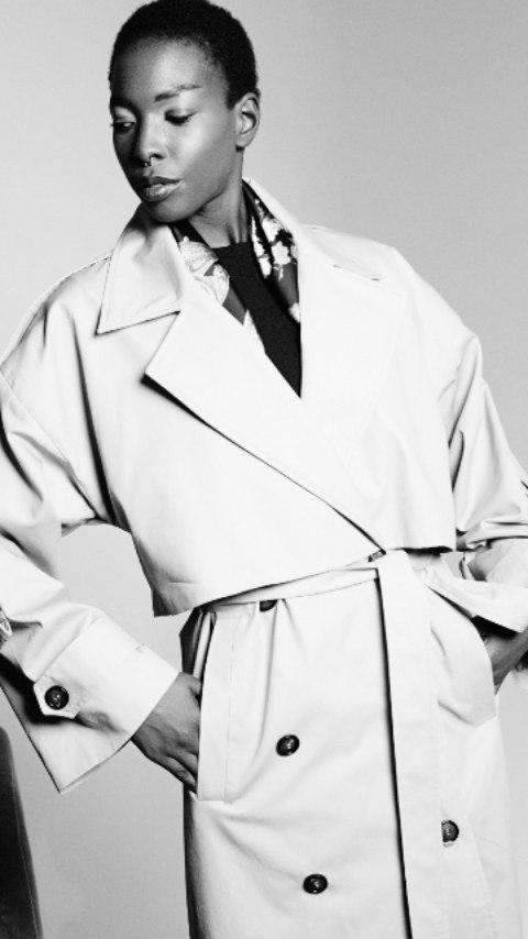 Femme portant un trench-coat beige avec des boutons, regardant sur le côté dans un style monochrome.