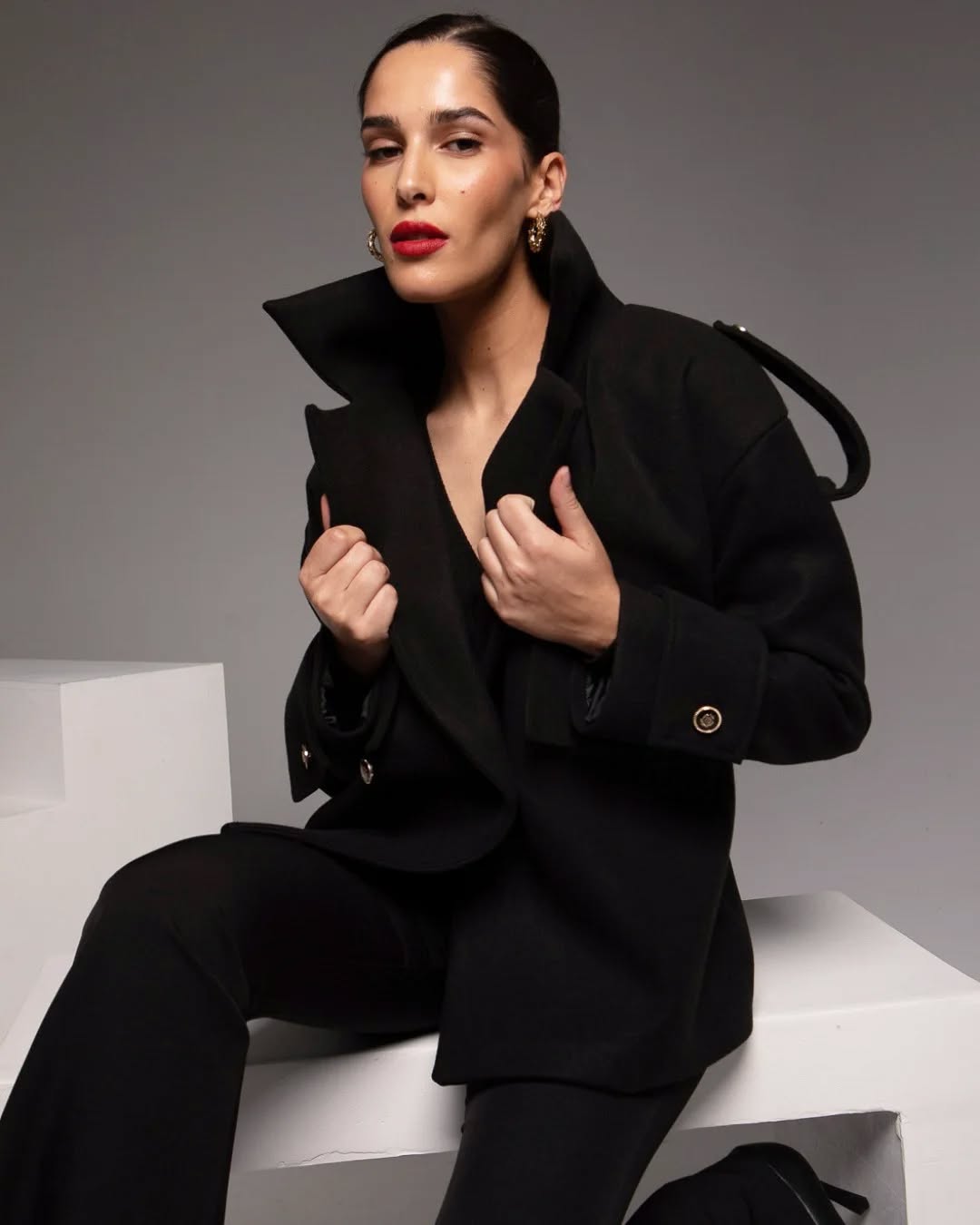 Femme portant un manteau noir avec un col relevé, posant avec une expression confiante et des accessoires dorés.