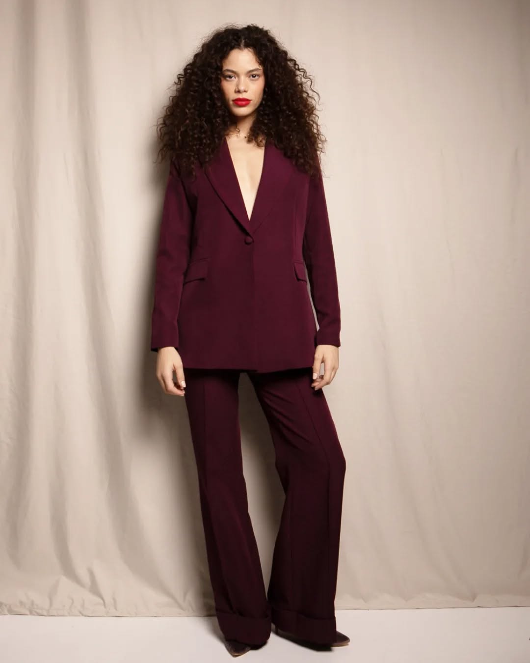 Femme portant un costume bordeaux avec un blazer à col en V et un pantalon assorti, cheveux bouclés et rouge à lèvres rouge.