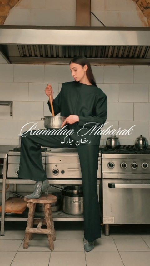 Femme en tenue verte remuant une casserole dans une cuisine professionnelle avec des équipements de cuisson.