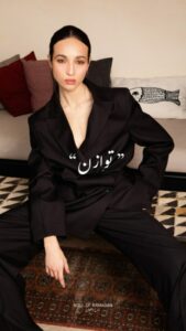 Femme en costume noir assise sur un canapé avec des coussins, portant un maquillage léger et une expression neutre.