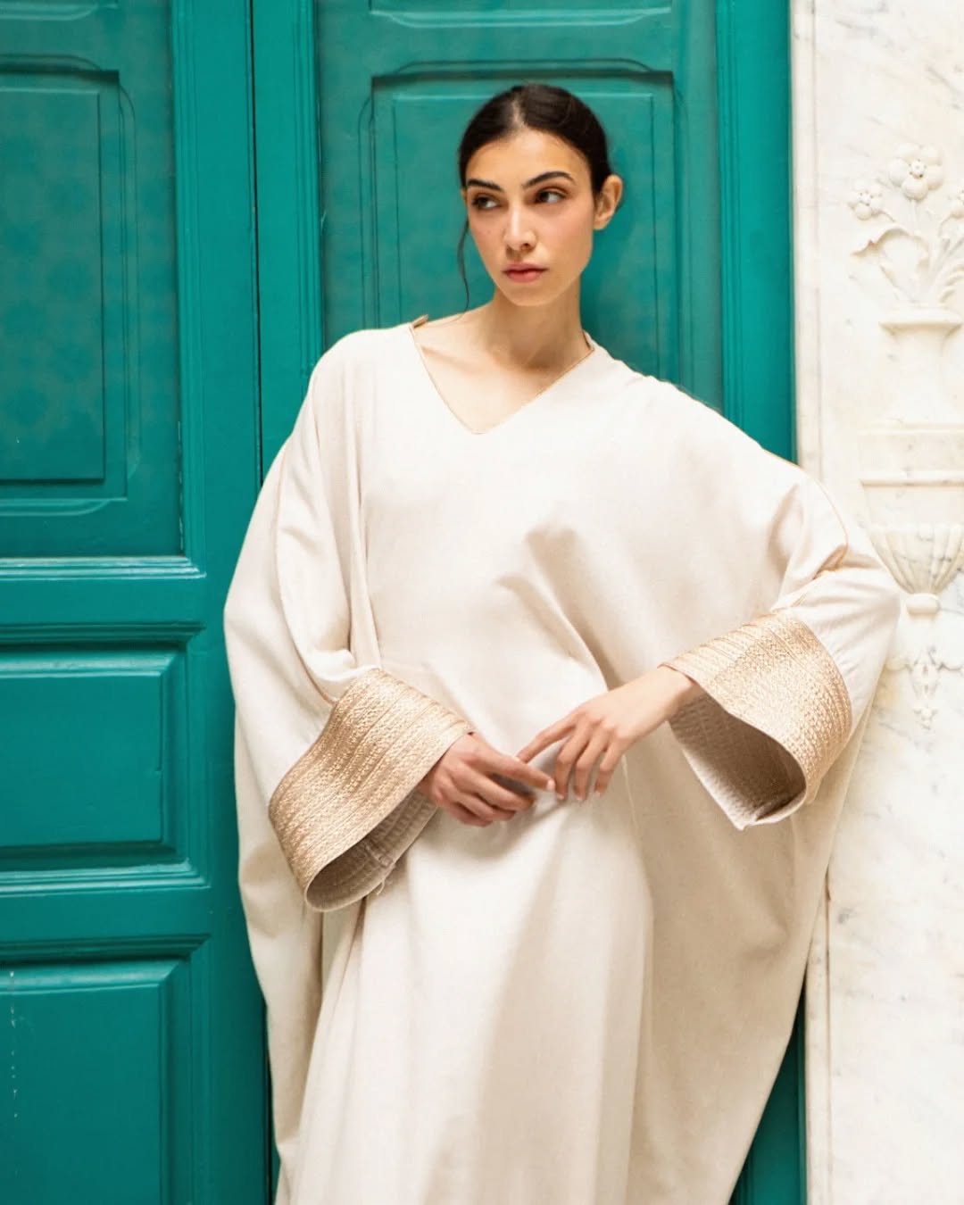 Femme portant une robe beige avec des manches larges et des détails en tissu texturé, posant devant une porte turquoise.