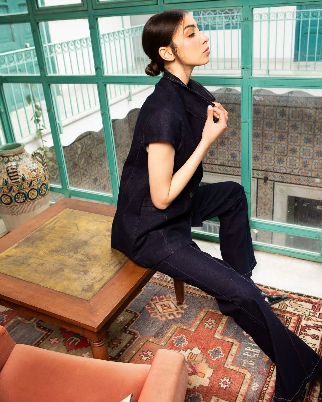 Femme portant un ensemble en denim bleu, assise sur une table en bois dans un intérieur lumineux avec des motifs décoratifs.