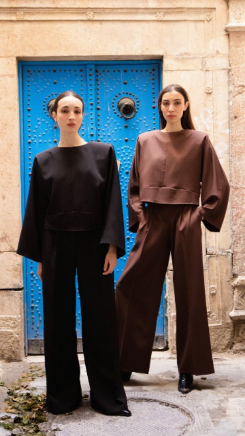 Deux silhouettes féminines portent des ensembles noirs et marron avec des manches amples devant une porte bleue ornée de rivets, sur un mur en pierre clair.