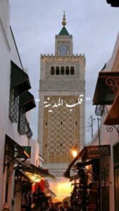 Minaret marocain en pierre gris clair dominant une rue du souk, façades blanches et motif de grille sur le mur, éclairage nocturne chaleureux.