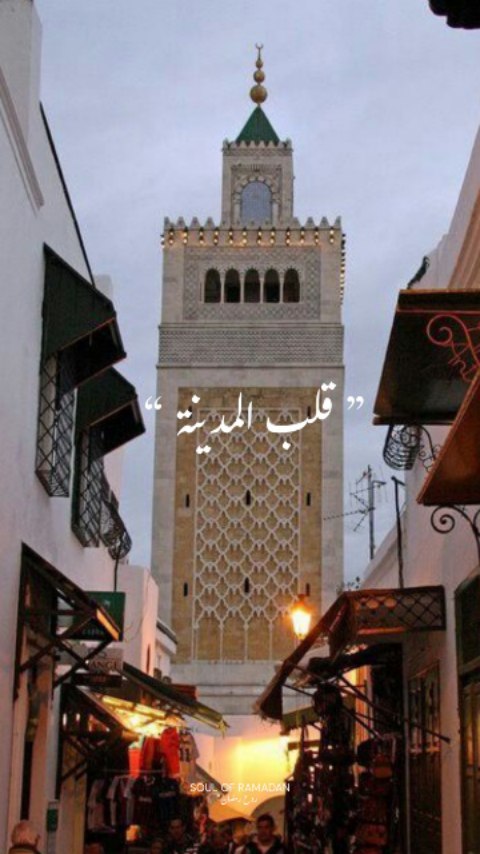 Minaret marocain en pierre gris clair dominant une rue du souk, façades blanches et motif de grille sur le mur, éclairage nocturne chaleureux.