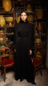 Femme vêtue d'une robe noire longue se tenant devant une étagère remplie de poteries jaunes et vertes, ambiance atelier, ambiance élégante et sobre