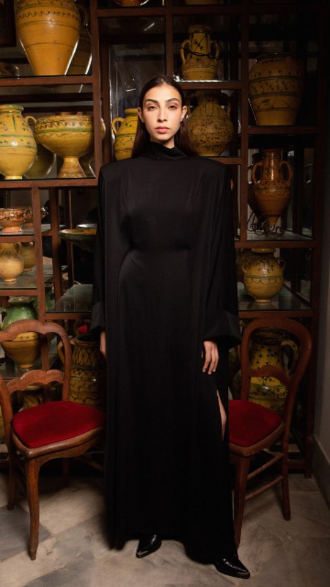 Femme vêtue d'une robe noire longue se tenant devant une étagère remplie de poteries jaunes et vertes, ambiance atelier, ambiance élégante et sobre