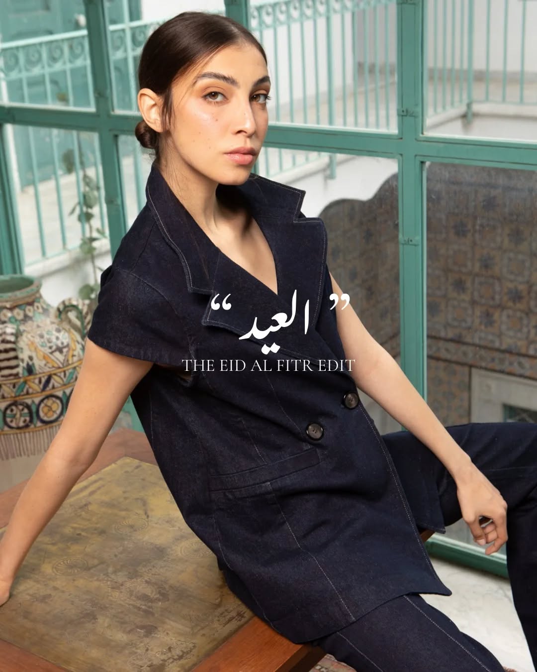 Portrait d'une jeune femme portant une tenue en denim bleu foncé, veste à col relevé et pantalon, assise près d'une véranda verte avec poterie décorative en arrière-plan.