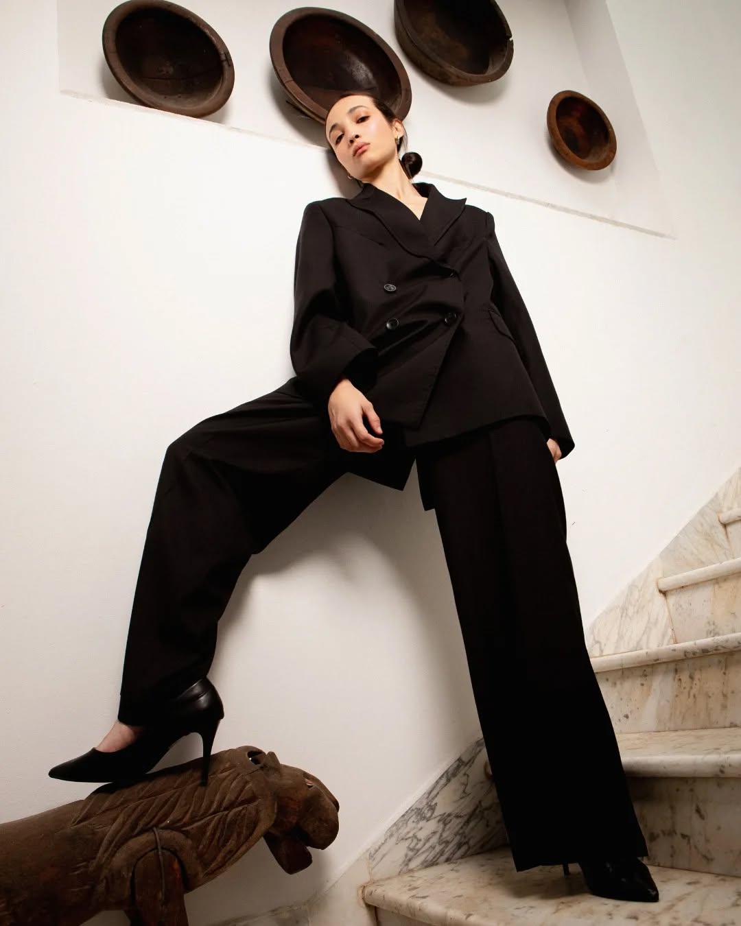 Modèle portant un ensemble noir sur mesure avec blazer croisé et pantalon large, talons noirs, dans un décor minimaliste avec des assiettes murales et un escalier en marbre.